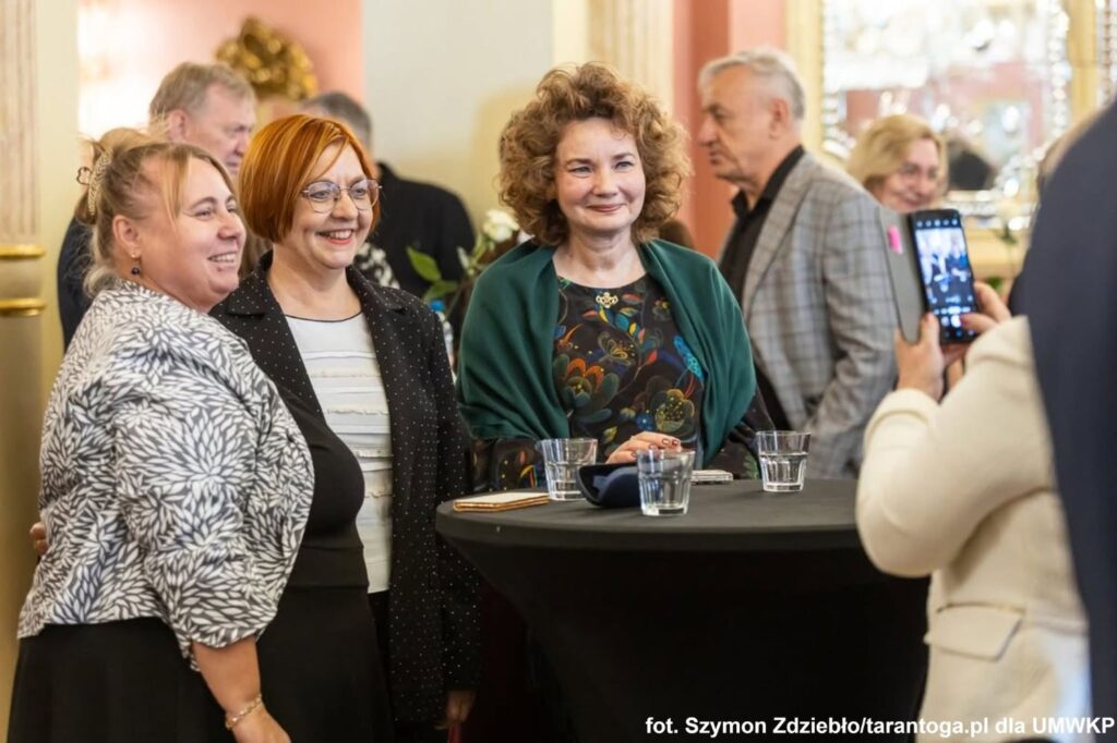 Trzy uśmiechnięte kobiety, stoją przy czarnym stoliku i pozują do zdjęcia,
