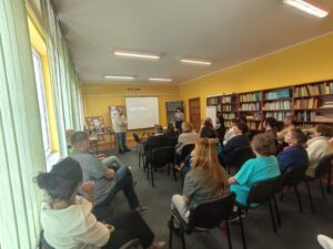 Sala biblioteczna pełna ludzi, przemawia do nich mężczyzna w jasnym swetrze.
