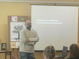 Mężczyzna w okularach i jasnym swetrze na tle ekranu.