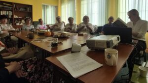 Kobiety przy stole - jedna czyta książkę, pozostałe słuchają.