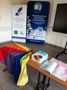 Na stole rozłożona chusta terapeutyczna, książki i kredki. W tle banery.
